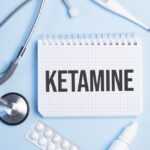 The word ketamine written on a white notepad on a blue background near a stethoscope, syringe, electronic thermometer and pills. Medical concept