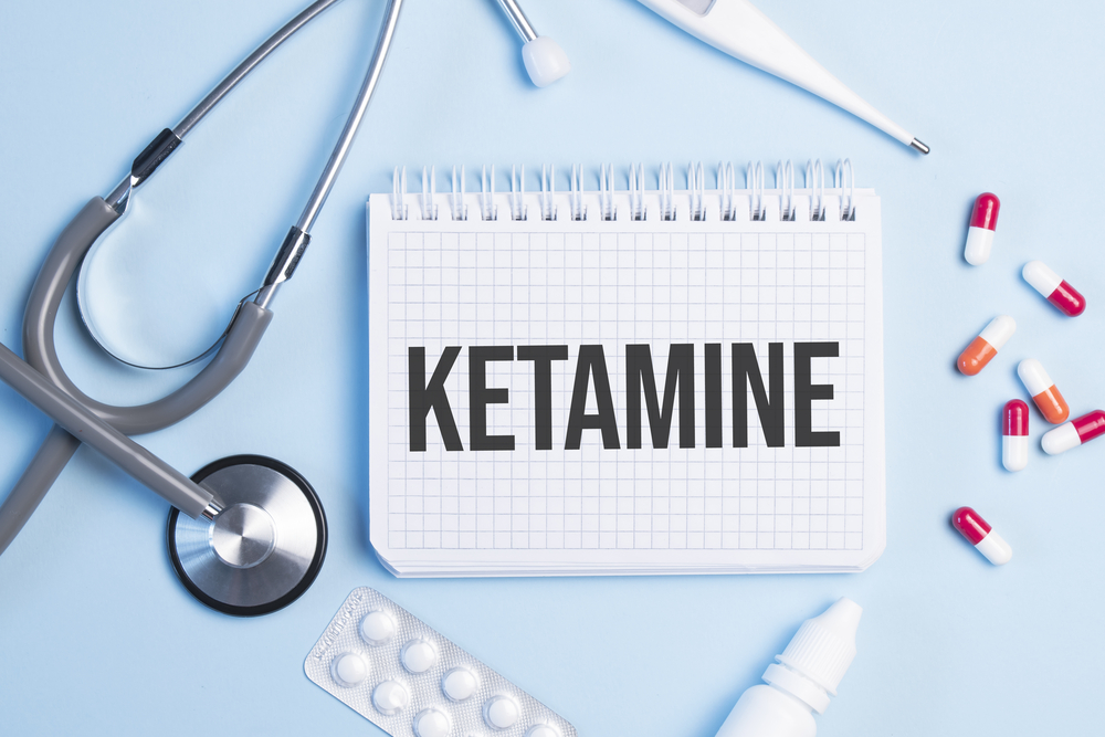 The word ketamine written on a white notepad on a blue background near a stethoscope, syringe, electronic thermometer and pills. Medical concept